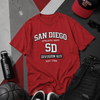 Red t-shirt with 'San Diego Athletic Dept' print, surrounded by accessories on a gray and red background.