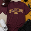 Maroon t-shirt with 'San Diego 619' text, white sneakers, black pants, sunglasses, and a cap on a patterned surface.
