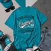 Turquoise t-shirt with 'San Diego Champs' text, surrounded by a cap, sunglasses, jeans, and sneakers on a gray surface.