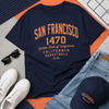 Navy t-shirt with 'San Francisco 1470' text, sunglasses, white sneakers, jeans, and a cap on a multicolored surface.