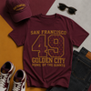 Maroon t-shirt with 'San Francisco 49 Golden City' text, maroon cap, sunglasses, and sneakers on a multicolored background.