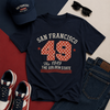 Navy t-shirt with 'San Francisco 49 The Golden State' design, paired with a cap, sunglasses, shoes, and jeans on a multicolored background.