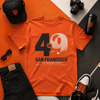 Orange t-shirt with 'San Francisco' design, black cap, camera, sunglasses, and shoes on a geometric patterned surface.