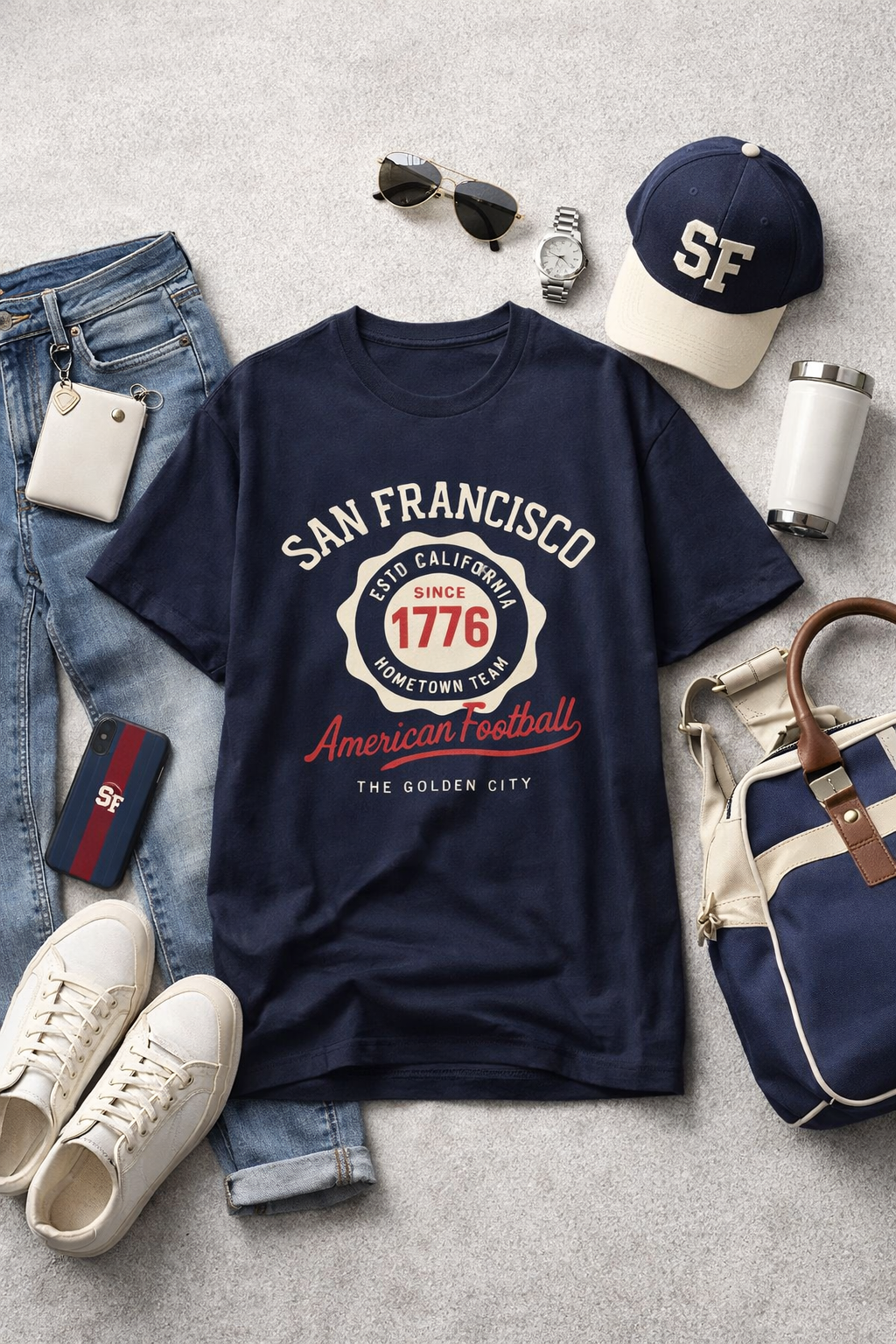 Navy blue t-shirt with 'San Francisco' text and graphics, surrounded by jeans, sneakers, a cap, sunglasses, a watch, a bag, and a phone on a gray surface.