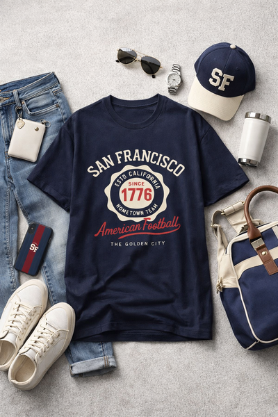 Navy blue t-shirt with 'San Francisco' text and graphics, surrounded by jeans, sneakers, a cap, sunglasses, a watch, a bag, and a phone on a gray surface.