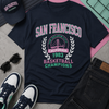 Navy blue t-shirt with 'San Francisco Basketball Champions' text, sunglasses, cap, and jeans on a textured surface.