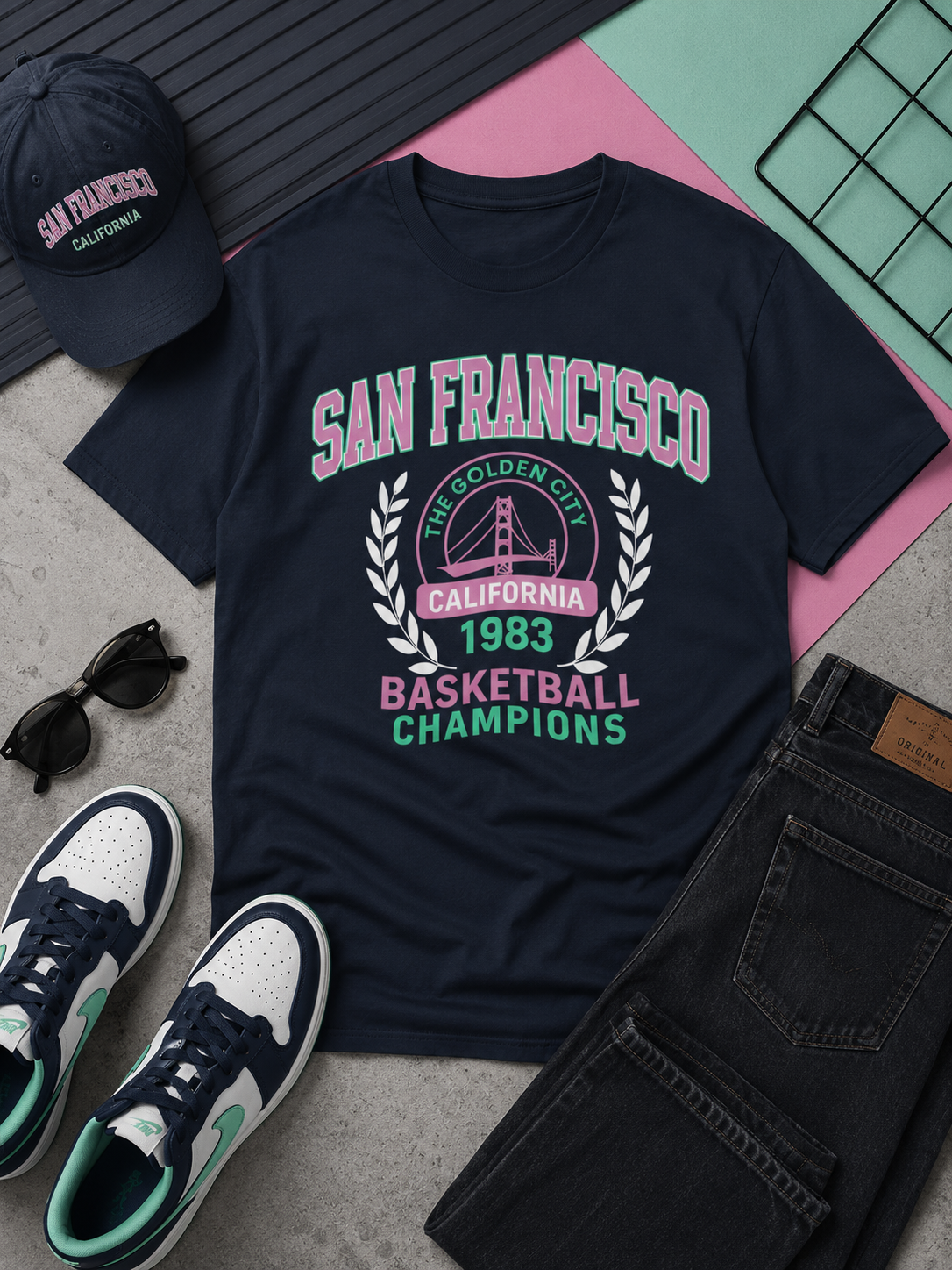 Navy blue t-shirt with 'San Francisco Basketball Champions' text, sunglasses, cap, and jeans on a textured surface.
