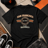Black t-shirt with 'San Francisco Baseball Club' design, surrounded by a cap, sneakers, and a baseball on a patterned surface.