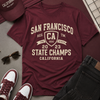 Maroon t-shirt with 'San Francisco State Champs' text, maroon cap, white sneakers, sunglasses, and black pants on a gray surface.