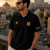 Man wearing a black striped polo shirt with a flag design, standing in an urban setting with buildings and traffic in the background.