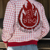 Person wearing a red and white checkered jacket with 'We've Got BBQ' text, standing in front of a 'Texas Style BBQ Low & Slow' sign.
